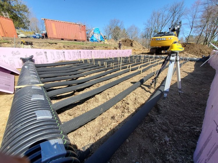 Construction site featuring a network of black piping laid out on the ground, surrounded by pink insulation boards. A yellow excavator is visible in the background, with shipping containers and workers present in the area. The scene showcases preparation for underground utility installation or drainage systems.