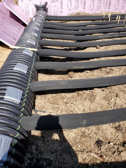 Installation of black piping laid out on a construction site, with insulation and supporting materials visible on the ground. The image showcases a series of parallel pipes, indicating preparation for underground utility work or geothermal heating systems.