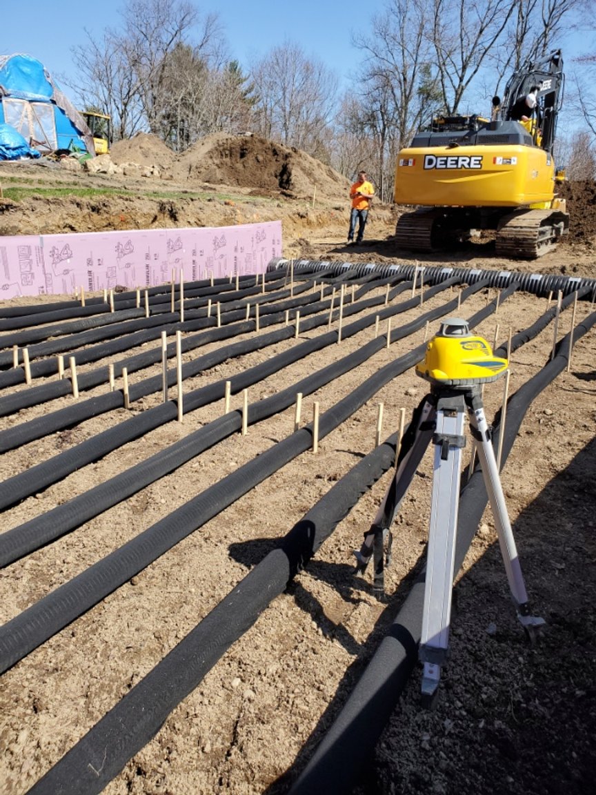 Construction site featuring a surveyor's tripod and equipment, with black drainage pipes laid out in rows. A yellow excavator is positioned nearby, and a worker in an orange shirt is overseeing the site. Pink insulation boards are visible in the background, indicating preparation for building foundations.