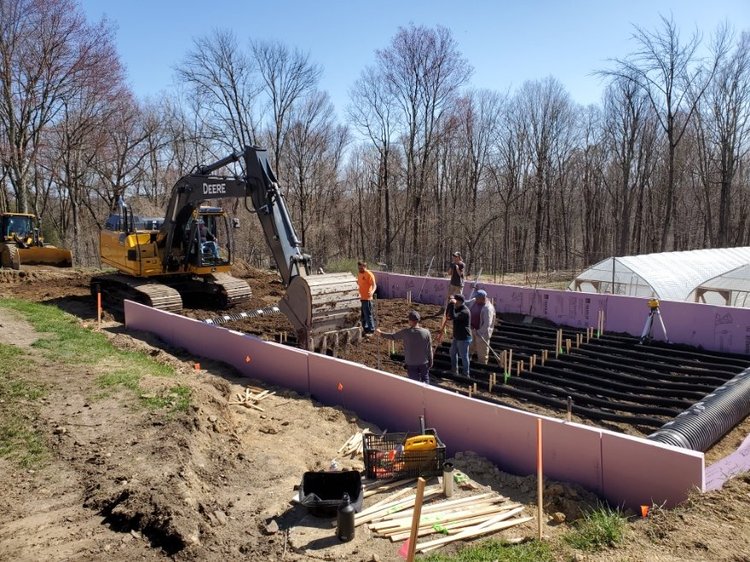 Construction site featuring a large excavator and several workers preparing the foundation for a building, with a pink insulated wall and rows of soil beds in the background, surrounded by trees and a greenhouse.