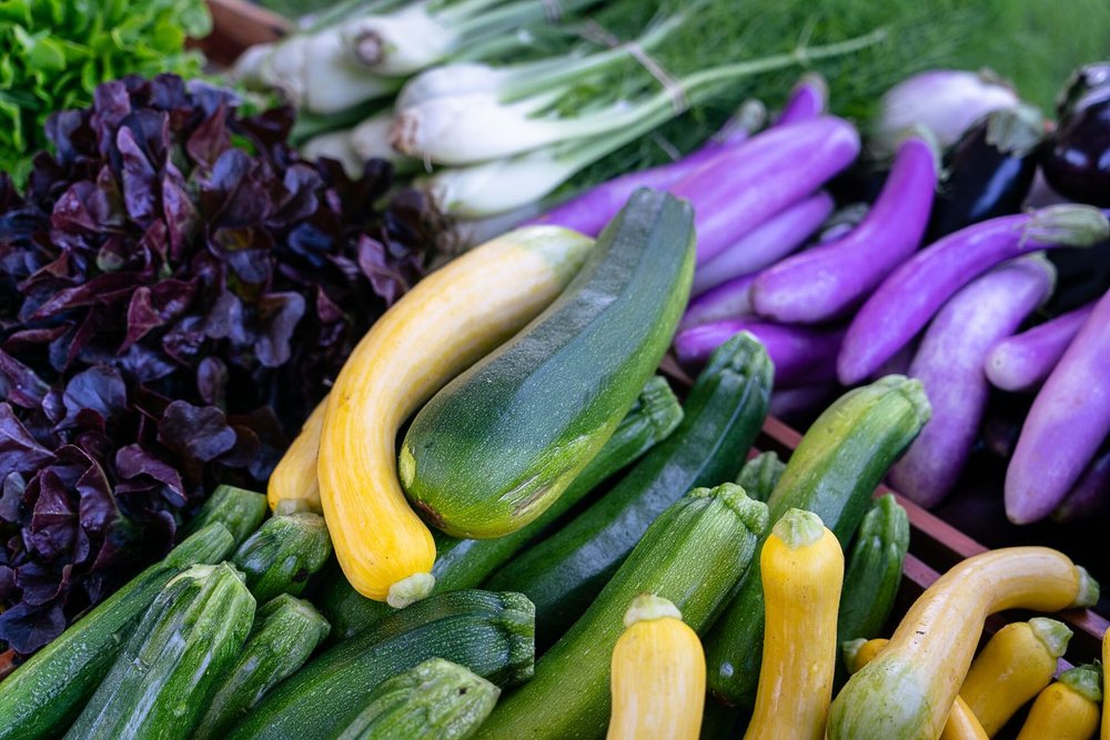 Fresh assortment of colorful vegetables including green and yellow zucchinis, purple eggplants, and red leaf lettuce, displayed at a local farmers market.